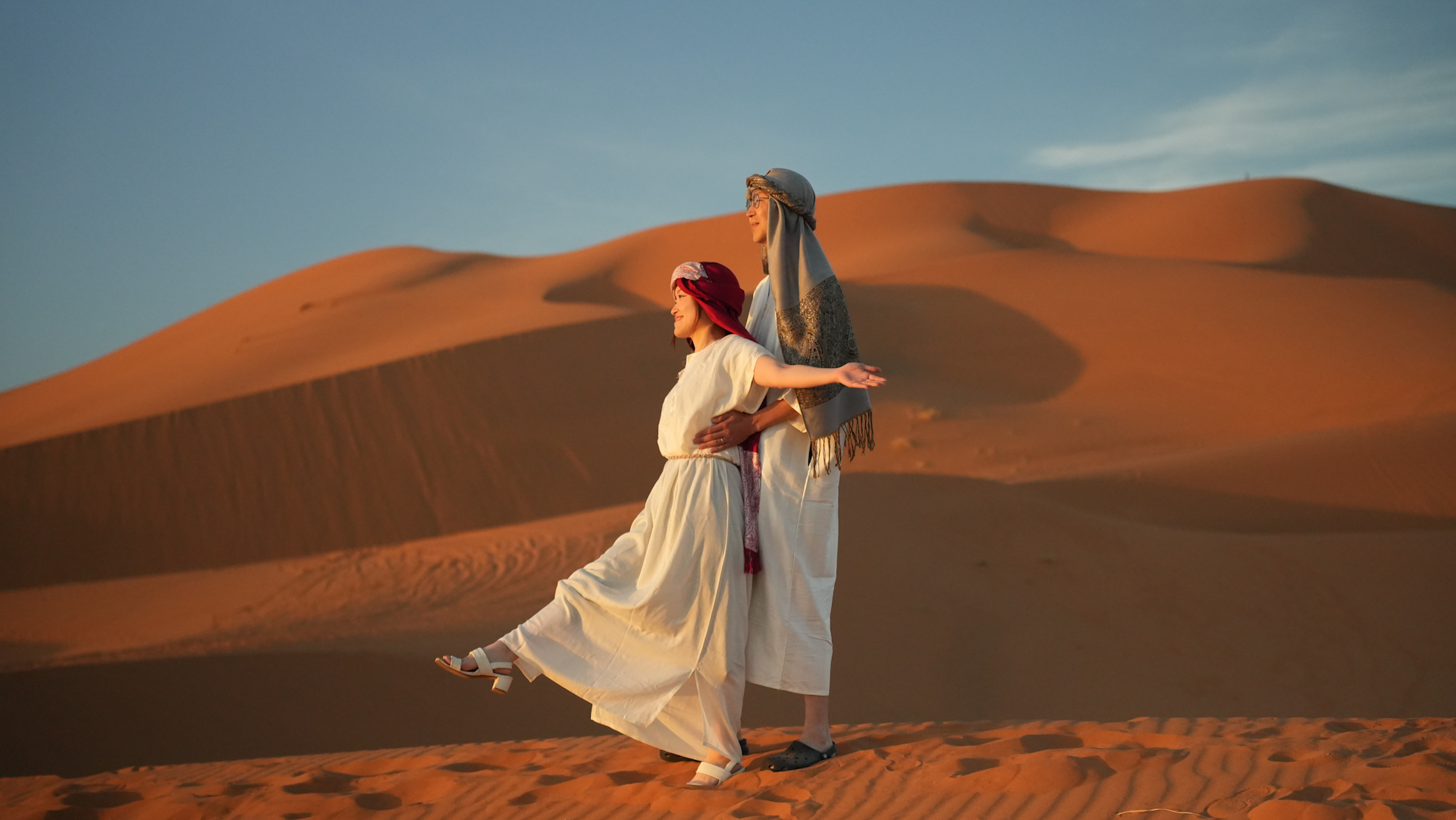 Couple standing together on golden sand dunes at sunset in Erg Chebbi Merzouga Sahara desert Morocco, captured during a professional Merzouga desert photoshoot by Inmerzouga photographer and drone services.